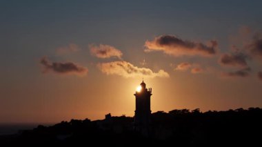 Cascais, Portekiz 'de dramatik hareket eden bulutları olan bir sahil uçurumundaki deniz fenerinin arkasındaki gün batımının sinematik arka plan görüntüsü.