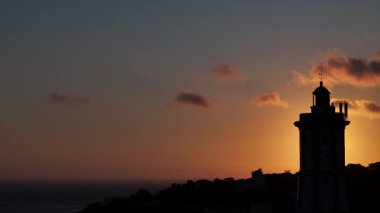 Cascais, Portekiz 'de dramatik hareket eden bulutları olan bir sahil uçurumundaki deniz fenerinin arkasındaki gün batımının sinematik arka plan görüntüsü.
