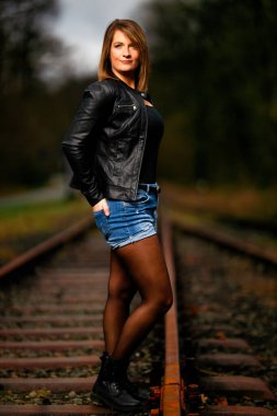 Deri ceketli genç bir kadın demiryolu raylarında poz veriyor, kameraya bakıyor ve olay yerini aydınlatıyor. Stüdyo ışığı gibi görün. Flash serbest bırakıldı.