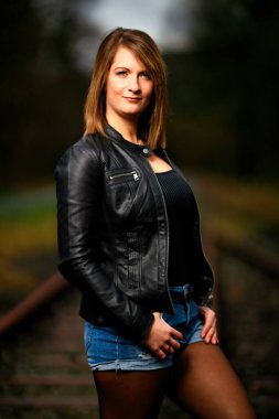 Deri ceketli genç bir kadın demiryolu raylarında poz veriyor, kameraya bakıyor ve olay yerini aydınlatıyor. Stüdyo ışığı gibi görün. Flash serbest bırakıldı.