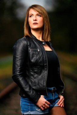 Deri ceketli genç bir kadın demiryolu raylarında poz veriyor, kameraya bakıyor ve olay yerini aydınlatıyor. Stüdyo ışığı gibi görün. Flash serbest bırakıldı.