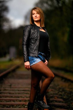 Deri ceketli genç bir kadın demiryolu raylarında poz veriyor, kameraya bakıyor ve olay yerini aydınlatıyor. Stüdyo ışığı gibi görün. Flash serbest bırakıldı.