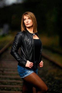 Deri ceketli genç bir kadın demiryolu raylarında poz veriyor, kameraya bakıyor ve olay yerini aydınlatıyor. Stüdyo ışığı gibi görün. Flash serbest bırakıldı.