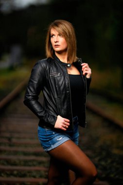 Deri ceketli genç bir kadın demiryolu raylarında poz veriyor, kameraya bakıyor ve olay yerini aydınlatıyor. Stüdyo ışığı gibi görün. Flash serbest bırakıldı.