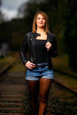 Deri ceketli genç bir kadın demiryolu raylarında poz veriyor, kameraya bakıyor ve olay yerini aydınlatıyor. Stüdyo ışığı gibi görün. Flash serbest bırakıldı.