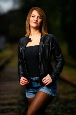 Deri ceketli genç bir kadın demiryolu raylarında poz veriyor, kameraya bakıyor ve olay yerini aydınlatıyor. Stüdyo ışığı gibi görün. Flash serbest bırakıldı.