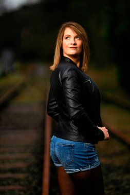Deri ceketli genç bir kadın demiryolu raylarında poz veriyor, kameraya bakıyor ve olay yerini aydınlatıyor. Stüdyo ışığı gibi görün. Flash serbest bırakıldı.