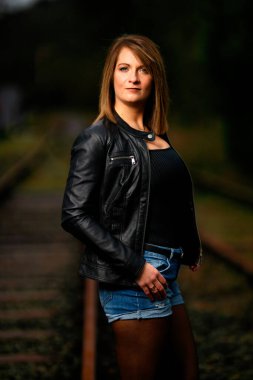 Deri ceketli genç bir kadın demiryolu raylarında poz veriyor, kameraya bakıyor ve olay yerini aydınlatıyor. Stüdyo ışığı gibi görün. Flash serbest bırakıldı.