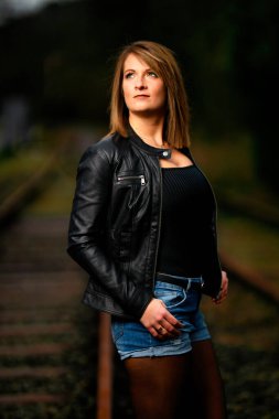 Deri ceketli genç bir kadın demiryolu raylarında poz veriyor, kameraya bakıyor ve olay yerini aydınlatıyor. Stüdyo ışığı gibi görün. Flash serbest bırakıldı.