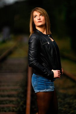 Deri ceketli genç bir kadın demiryolu raylarında poz veriyor, kameraya bakıyor ve olay yerini aydınlatıyor. Stüdyo ışığı gibi görün. Flash serbest bırakıldı.
