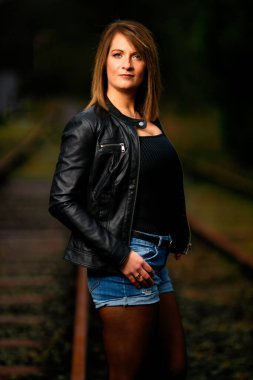Deri ceketli genç bir kadın demiryolu raylarında poz veriyor, kameraya bakıyor ve olay yerini aydınlatıyor. Stüdyo ışığı gibi görün. Flash serbest bırakıldı.