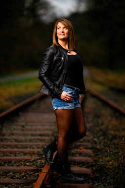 Deri ceketli genç bir kadın demiryolu raylarında poz veriyor, kameraya bakıyor ve olay yerini aydınlatıyor. Stüdyo ışığı gibi görün. Flash serbest bırakıldı.