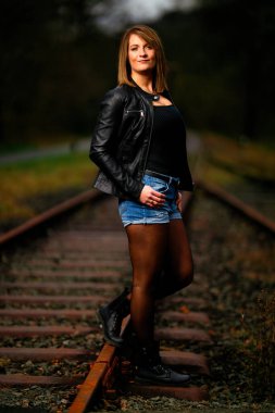 Deri ceketli genç bir kadın demiryolu raylarında poz veriyor, kameraya bakıyor ve olay yerini aydınlatıyor. Stüdyo ışığı gibi görün. Flash serbest bırakıldı.