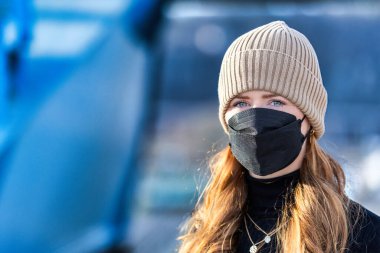 Resim kızıl saçlı genç bir kadını gösteriyor. Kendini bulaşıcı hastalıklardan korumak için FFP 2 maskesi takıyor. Saçları uzun ve baştan çıkarıcı, örgü şapka takıyor..