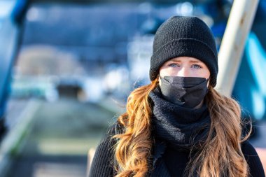 Resim kızıl saçlı genç bir kadını gösteriyor. Kendini bulaşıcı hastalıklardan korumak için FFP 2 maskesi takıyor. Saçları uzun ve baştan çıkarıcı, örgü şapka takıyor..