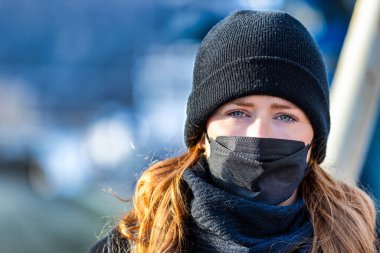 Resim kızıl saçlı genç bir kadını gösteriyor. Kendini bulaşıcı hastalıklardan korumak için FFP 2 maskesi takıyor. Saçları uzun ve baştan çıkarıcı, örgü şapka takıyor..