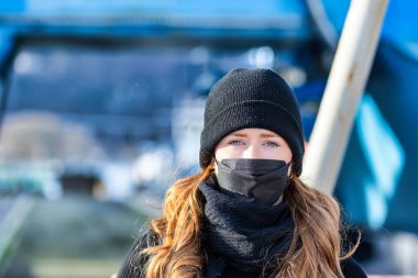 Resim kızıl saçlı genç bir kadını gösteriyor. Kendini bulaşıcı hastalıklardan korumak için FFP 2 maskesi takıyor. Saçları uzun ve baştan çıkarıcı, örgü şapka takıyor..