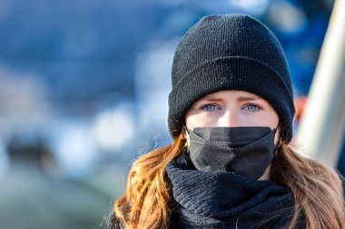Resim kızıl saçlı genç bir kadını gösteriyor. Kendini bulaşıcı hastalıklardan korumak için FFP 2 maskesi takıyor. Saçları uzun ve baştan çıkarıcı, örgü şapka takıyor..
