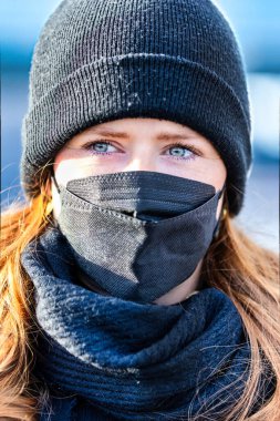Resim kızıl saçlı genç bir kadını gösteriyor. Kendini bulaşıcı hastalıklardan korumak için FFP 2 maskesi takıyor. Saçları uzun ve baştan çıkarıcı, örgü şapka takıyor..