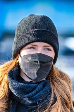 Resim kızıl saçlı genç bir kadını gösteriyor. Kendini bulaşıcı hastalıklardan korumak için FFP 2 maskesi takıyor. Saçları uzun ve baştan çıkarıcı, örgü şapka takıyor..