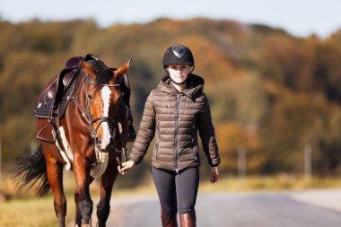 Genç bir kadın güneşli bir sonbahar gününde yolda yürüyor ve atını gezintiye çıkarıyor. Binicilik kıyafetleri ve eyerli bir atla. Arka planda sonbahar ormanı olan manzara formatında fotoğraf.