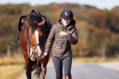 Genç bir kadın güneşli bir sonbahar gününde yolda yürüyor ve atını gezintiye çıkarıyor. Binicilik kıyafetleri ve eyerli bir atla. Arka planda sonbahar ormanı olan manzara formatında fotoğraf.