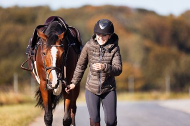 Genç bir kadın güneşli bir sonbahar gününde yolda yürüyor ve atını gezintiye çıkarıyor. Binicilik kıyafetleri ve eyerli bir atla. Arka planda sonbahar ormanı olan manzara formatında fotoğraf.