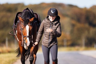 Genç bir kadın güneşli bir sonbahar gününde yolda yürüyor ve atını gezintiye çıkarıyor. Binicilik kıyafetleri ve eyerli bir atla. Arka planda sonbahar ormanı olan manzara formatında fotoğraf.