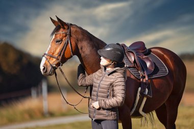 Sonbahar doğasında, gün ışığında yanında binicisi olan bir çayırdaki at..