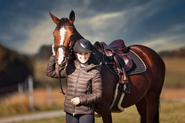 Sonbahar doğasında, gün ışığında yanında binicisi olan bir çayırdaki at..
