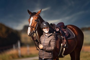Sonbahar doğasında, gün ışığında yanında binicisi olan bir çayırdaki at..