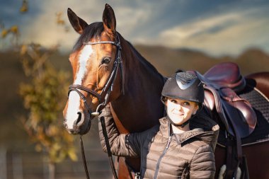 Sonbahar doğasında, gün ışığında yanında binicisi olan bir çayırdaki at..