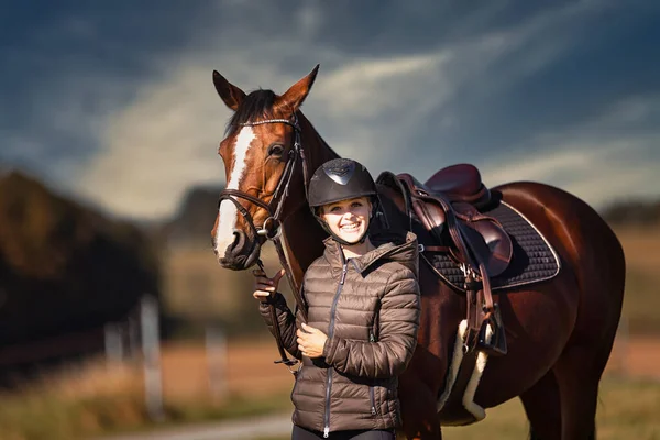 Sonbahar doğasında, gün ışığında yanında binicisi olan bir çayırdaki at..