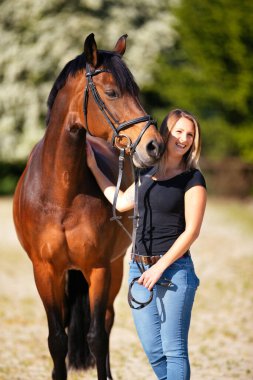 Genç bir kadın atıyla güneşli bir arenada duruyor. Arka planda, çiçek açan ağaçlar ve yeşil doğa, bulanık, kahverengi at ve kot pantolon giymiş kadının önünde izole edilmiş model..