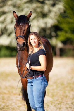 Genç bir kadın atıyla güneşli bir arenada duruyor. Arka planda, çiçek açan ağaçlar ve yeşil doğa, bulanık, kahverengi at ve kot pantolon giymiş kadının önünde izole edilmiş model..
