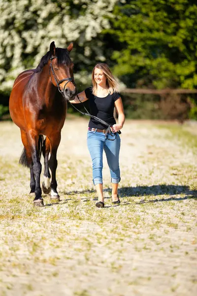 Genç bir kadın atıyla güneşli bir arenada duruyor. Arka planda, çiçek açan ağaçlar ve yeşil doğa, bulanık, kahverengi at ve kot pantolon giymiş kadının önünde izole edilmiş model..