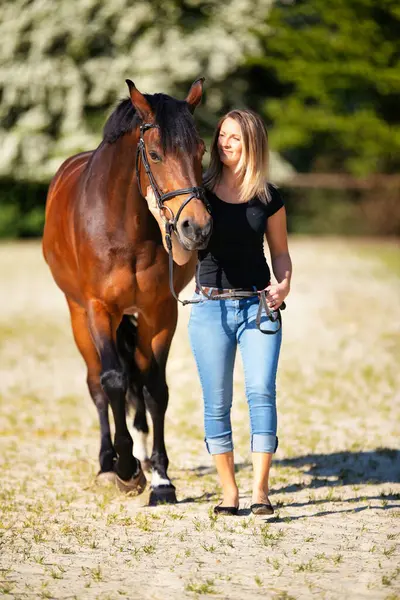 Genç bir kadın atıyla güneşli bir arenada duruyor. Arka planda, çiçek açan ağaçlar ve yeşil doğa, bulanık, kahverengi at ve kot pantolon giymiş kadının önünde izole edilmiş model..