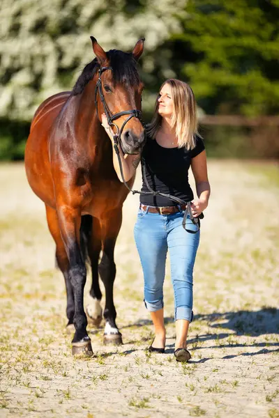 Genç bir kadın atıyla güneşli bir arenada duruyor. Arka planda, çiçek açan ağaçlar ve yeşil doğa, bulanık, kahverengi at ve kot pantolon giymiş kadının önünde izole edilmiş model..