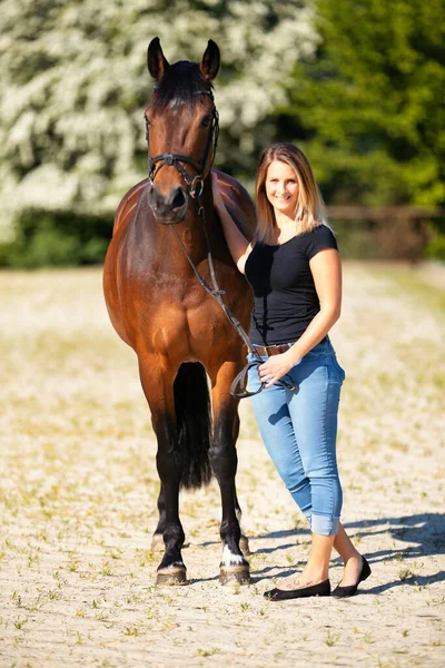 Genç bir kadın atıyla güneşli bir arenada duruyor. Arka planda, çiçek açan ağaçlar ve yeşil doğa, bulanık, kahverengi at ve kot pantolon giymiş kadının önünde izole edilmiş model..