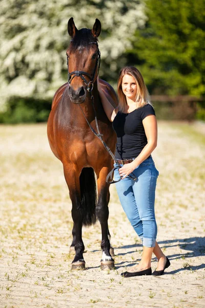 Genç bir kadın atıyla güneşli bir arenada duruyor. Arka planda, çiçek açan ağaçlar ve yeşil doğa, bulanık, kahverengi at ve kot pantolon giymiş kadının önünde izole edilmiş model..