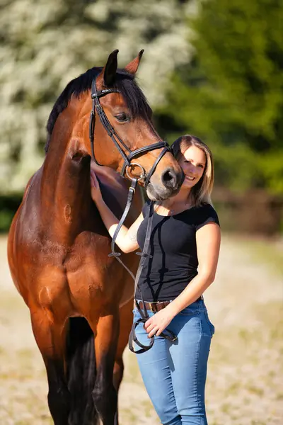 Genç bir kadın atıyla güneşli bir arenada duruyor. Arka planda, çiçek açan ağaçlar ve yeşil doğa, bulanık, kahverengi at ve kot pantolon giymiş kadının önünde izole edilmiş model..