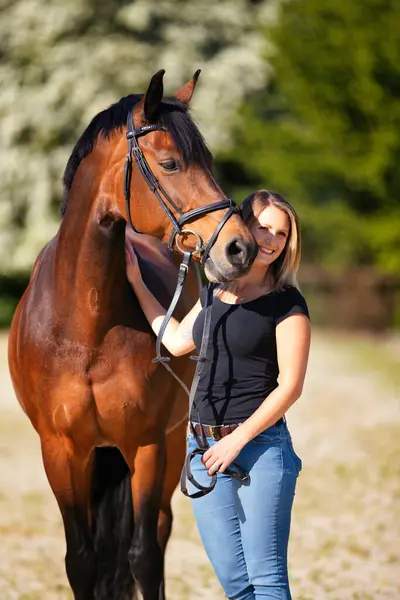 Genç bir kadın atıyla güneşli bir arenada duruyor. Arka planda, çiçek açan ağaçlar ve yeşil doğa, bulanık, kahverengi at ve kot pantolon giymiş kadının önünde izole edilmiş model..