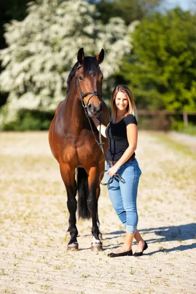 Genç bir kadın atıyla güneşli bir arenada duruyor. Arka planda, çiçek açan ağaçlar ve yeşil doğa, bulanık, kahverengi at ve kot pantolon giymiş kadının önünde izole edilmiş model..