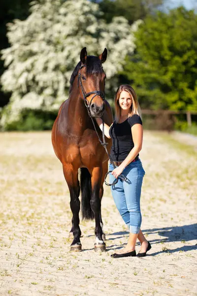 Genç bir kadın atıyla güneşli bir arenada duruyor. Arka planda, çiçek açan ağaçlar ve yeşil doğa, bulanık, kahverengi at ve kot pantolon giymiş kadının önünde izole edilmiş model..
