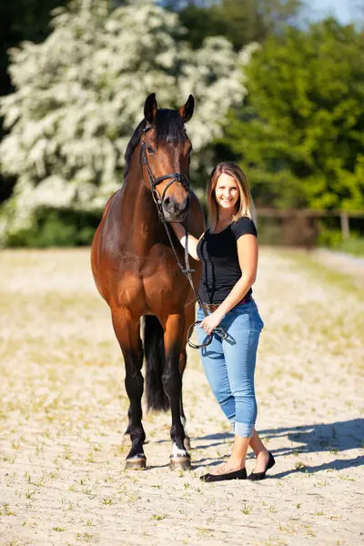 Genç bir kadın atıyla güneşli bir arenada duruyor. Arka planda, çiçek açan ağaçlar ve yeşil doğa, bulanık, kahverengi at ve kot pantolon giymiş kadının önünde izole edilmiş model..