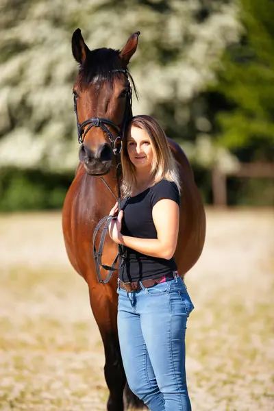 Genç bir kadın atıyla güneşli bir arenada duruyor. Arka planda, çiçek açan ağaçlar ve yeşil doğa, bulanık, kahverengi at ve kot pantolon giymiş kadının önünde izole edilmiş model..