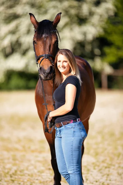 Genç bir kadın atıyla güneşli bir arenada duruyor. Arka planda, çiçek açan ağaçlar ve yeşil doğa, bulanık, kahverengi at ve kot pantolon giymiş kadının önünde izole edilmiş model..