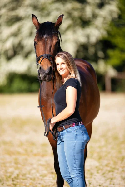 Genç bir kadın atıyla güneşli bir arenada duruyor. Arka planda, çiçek açan ağaçlar ve yeşil doğa, bulanık, kahverengi at ve kot pantolon giymiş kadının önünde izole edilmiş model..