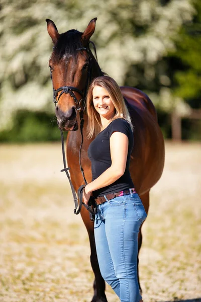 Genç bir kadın atıyla güneşli bir arenada duruyor. Arka planda, çiçek açan ağaçlar ve yeşil doğa, bulanık, kahverengi at ve kot pantolon giymiş kadının önünde izole edilmiş model..