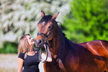 Genç bir kadın sıcak bir bahar gününün güneşinde atıyla portrede dikiliyor ve arka planda çiçek açan bir dikenli çalı var. Metin için boşluk olan manzara biçiminde resim.